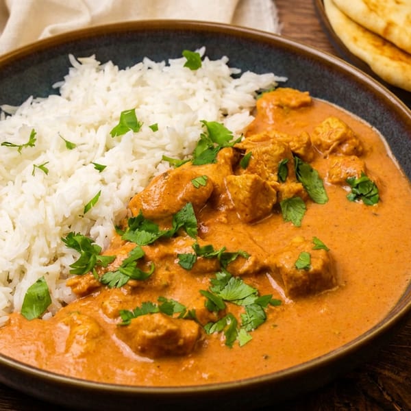 Chicken curry with white rice and cilantro in a bowl, with naan bread on a wooden table.
